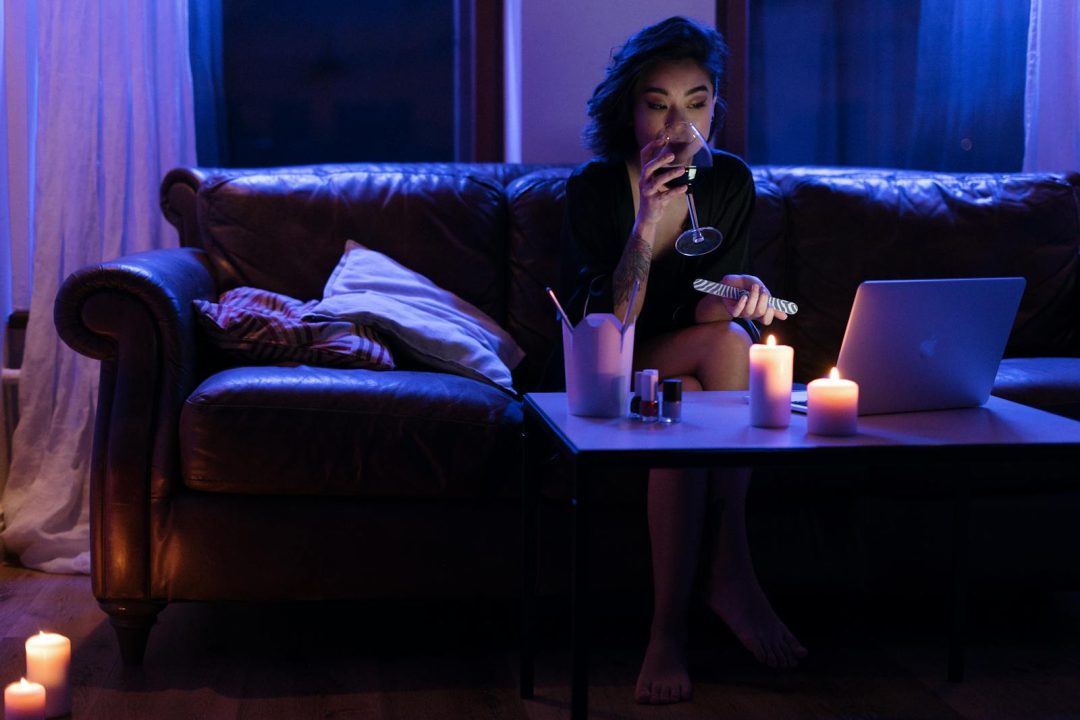 Woman relaxing with wine and laptop on a cozy evening illuminated by candles.